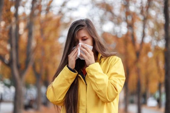 Young woman standing in a park wearing a yellow coat, holding a tissue to her nose while sneezing.
