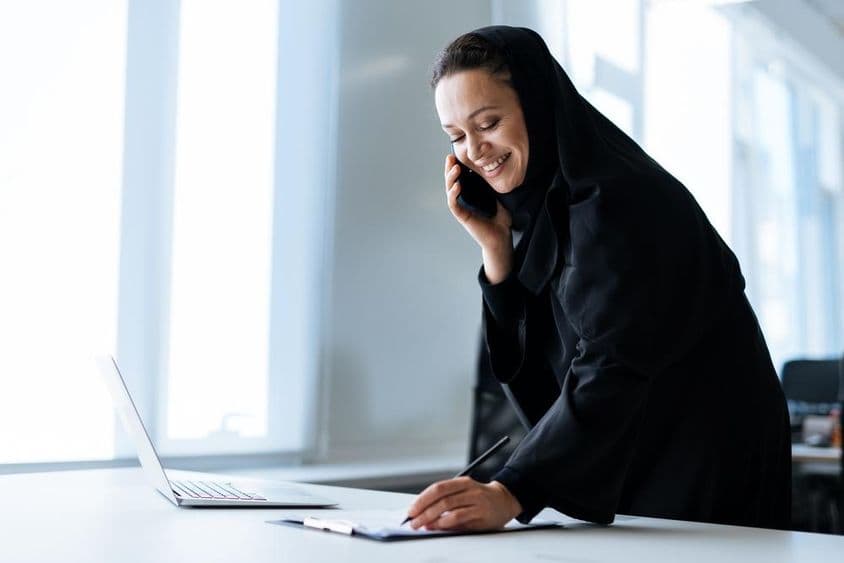 A beautiful woman in an abaya working on her computer while on the phone.