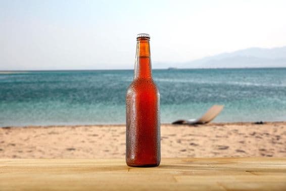 A bottle of cold beer on a wooden board table on a sandy beach shoreline.