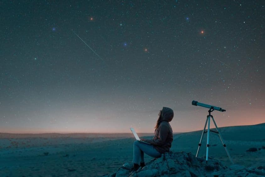 Woman sitting in the desert with a laptop next to a telescope at night, watching the starry sky.