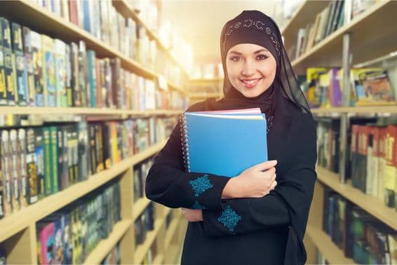 Arab student standing in library with books in arm.