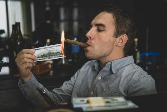 Young handsome man lighting cigar with hundred-dollar bill in a bar.