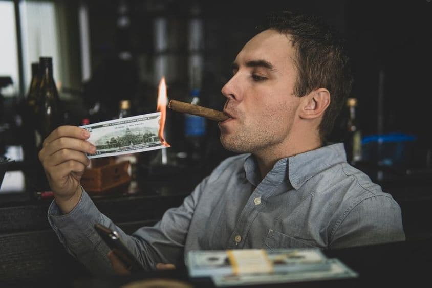 Young handsome man lighting cigar with hundred-dollar bill in a bar.