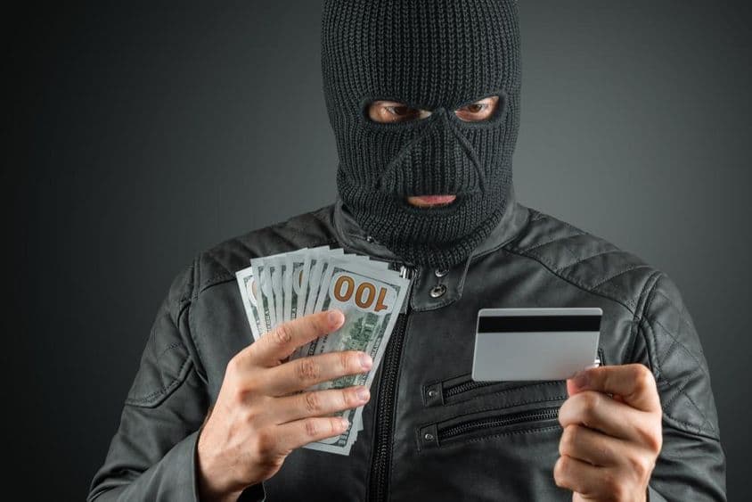 Criminal in ski mask holding credit card against a dark background.
