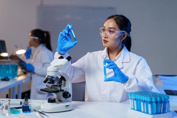 Asian female scientist in lab coat and gloves comparing two test tubes.