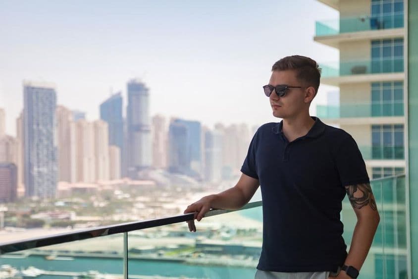 A young man in sunglasses standing on a skyscraper balcony overlooking Dubai.