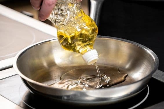 A hand pouring golden cooking oil from a plastic bottle into a stainless steel pan.