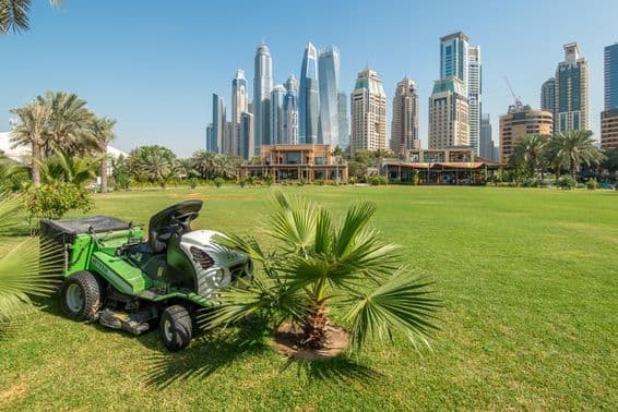 Lawn mower tractor on a green field in front of Dubai Marina skyscrapers.