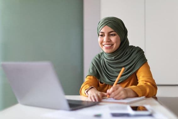 Young Muslim woman watching a webinar at home on a laptop and taking notes.