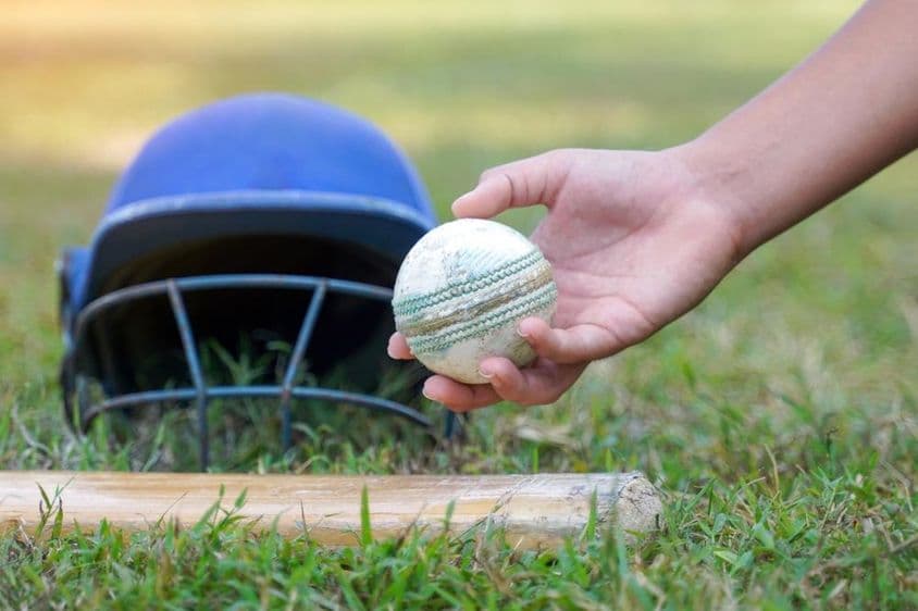 A player's hand picking up a cricket ball from among other cricket gear lying on the grass.