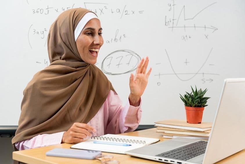 Female teacher in a hijab waving to students during an online class.