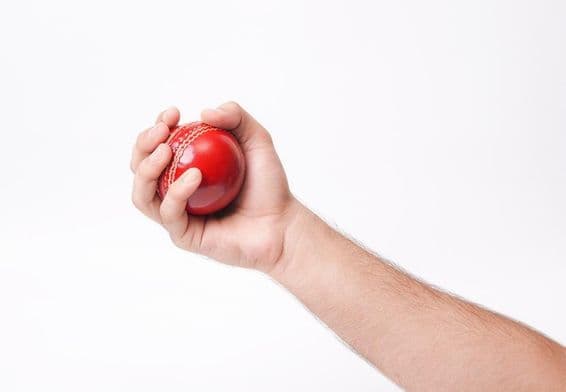 A male cricketer gripping a red cricket ball firmly.
