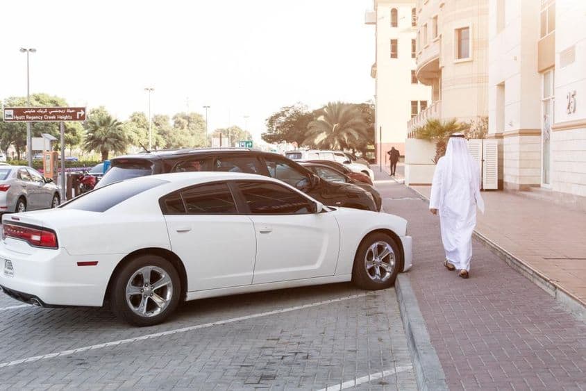 A man in traditional attire and a white Dodge in a parking lot.