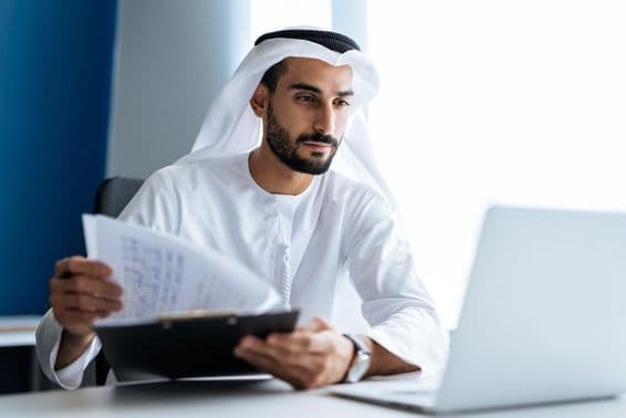 Man working with a laptop in Dubai office, wearing traditional white Emirati attire.