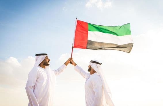 Arab men wearing kanduras walking in the desert holding a flag.