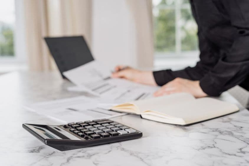 A woman in black clothing calculating expenses with a laptop.