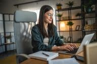 Adult woman working on a laptop and using a notebook in a home office.