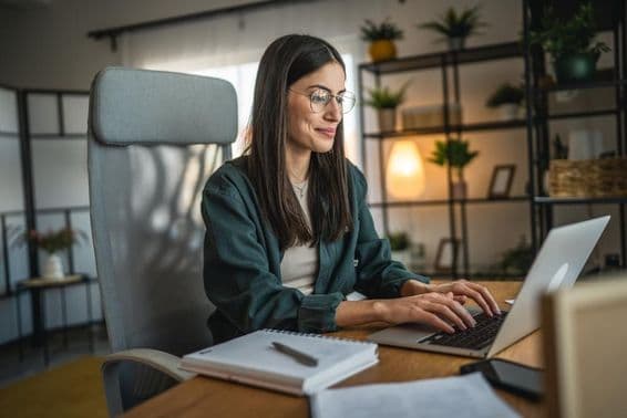 Adult woman working on a laptop and using a notebook in a home office.