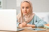 Little Muslim girl in hijab doing homework at home.