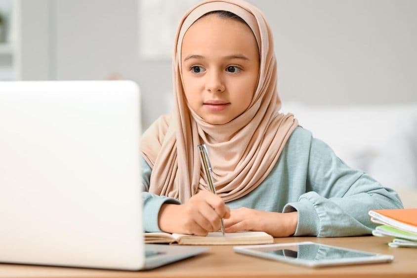 Little Muslim girl in hijab doing homework at home.
