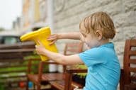 Soaked preschool boy pouring water out of a yellow rubber boot.
