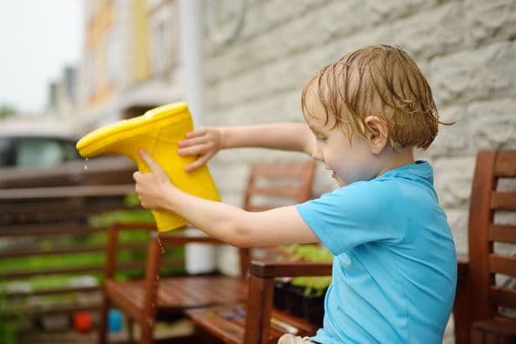 Soaked preschool boy pouring water out of a yellow rubber boot.
