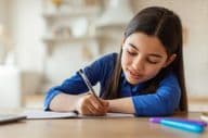 Arab schoolgirl taking notes while sitting at a table with a laptop at home.