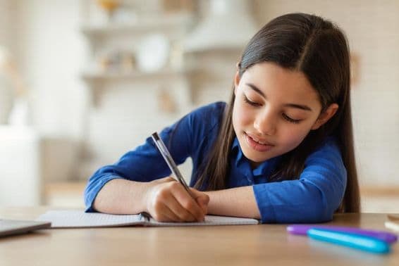 Arab schoolgirl taking notes while sitting at a table with a laptop at home.