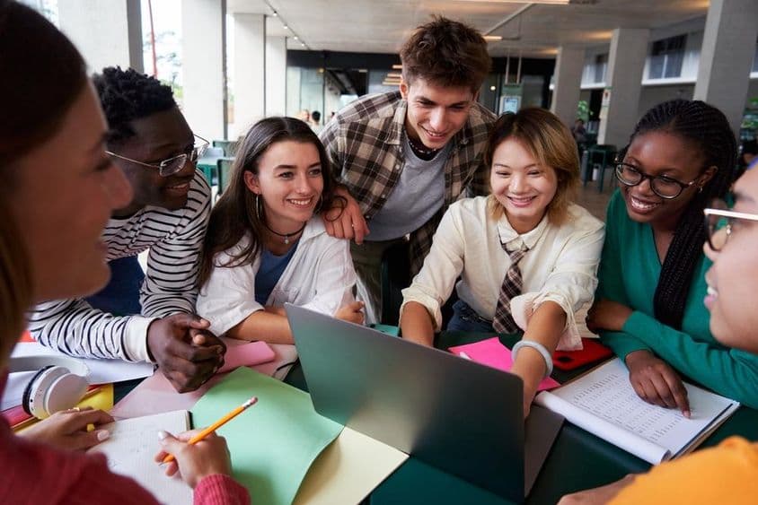 A cheerful group of university students sit at a table looking at a notebook.