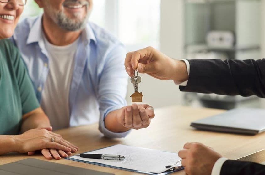 A real estate agent handing keys to a smiling elderly couple.