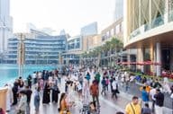 A diverse crowd of tourists and locals walks along the busy waterfront promenade in front of Dubai Mall.
