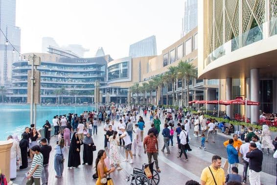 A diverse crowd of tourists and locals walks along the busy waterfront promenade in front of Dubai Mall.